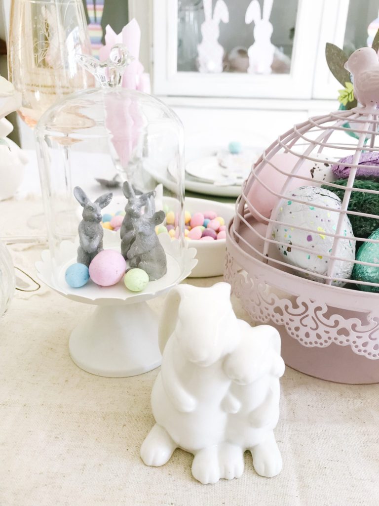 Three grey bunnies with speckled eggs in small glass cloche with pink bird cage and white ceramic buniies