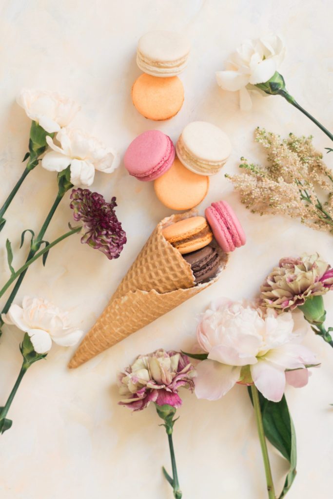 Pink, orange and cream macarons spilling out of an ice-cream cone while surrounded by flowers