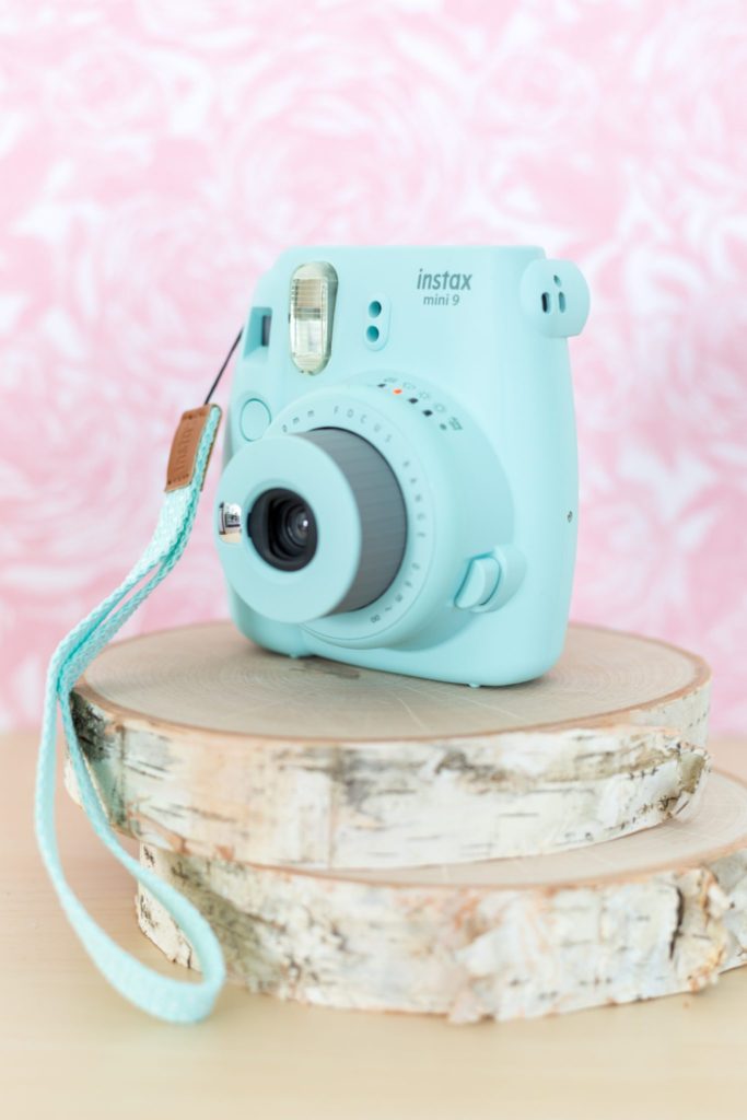 A turquoise Instax camera on top of two wooden logs with a pretty pink background